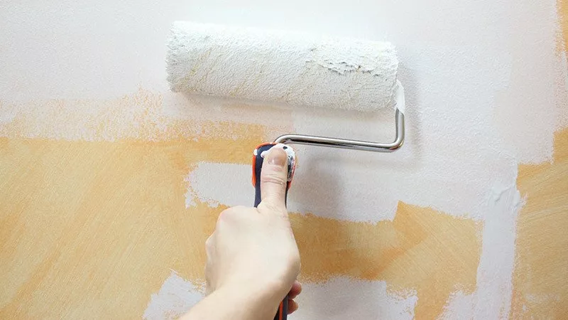 hand with paint roller painting white paint over a yellow indoor wall