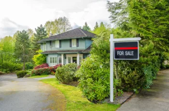 Green two-story home with for sale sign with lots of trees and manicured yard.