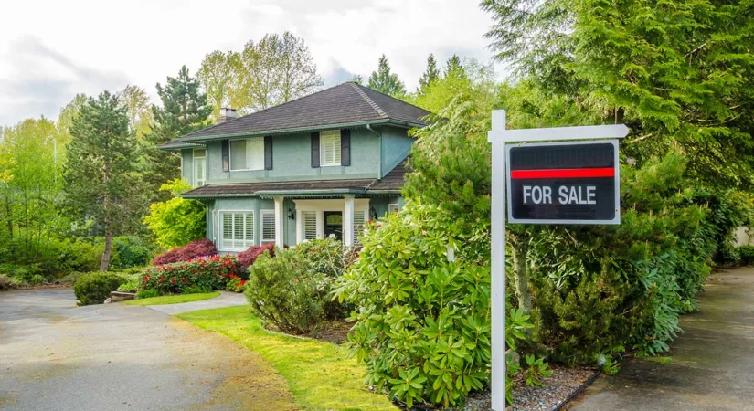 Green two-story home with for sale sign with lots of trees and manicured yard.