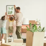 Man and two children hanging picture in new home, with unpacked boxes in foreground.