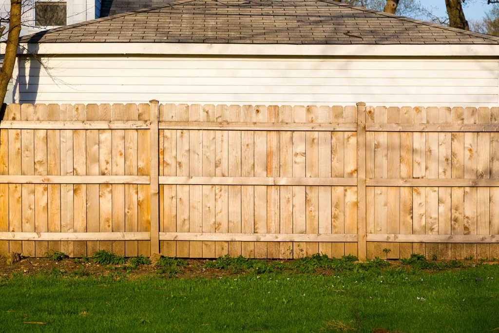New fence with unpainted or stained boards that touch the ground, with garage wall and roof behind. 