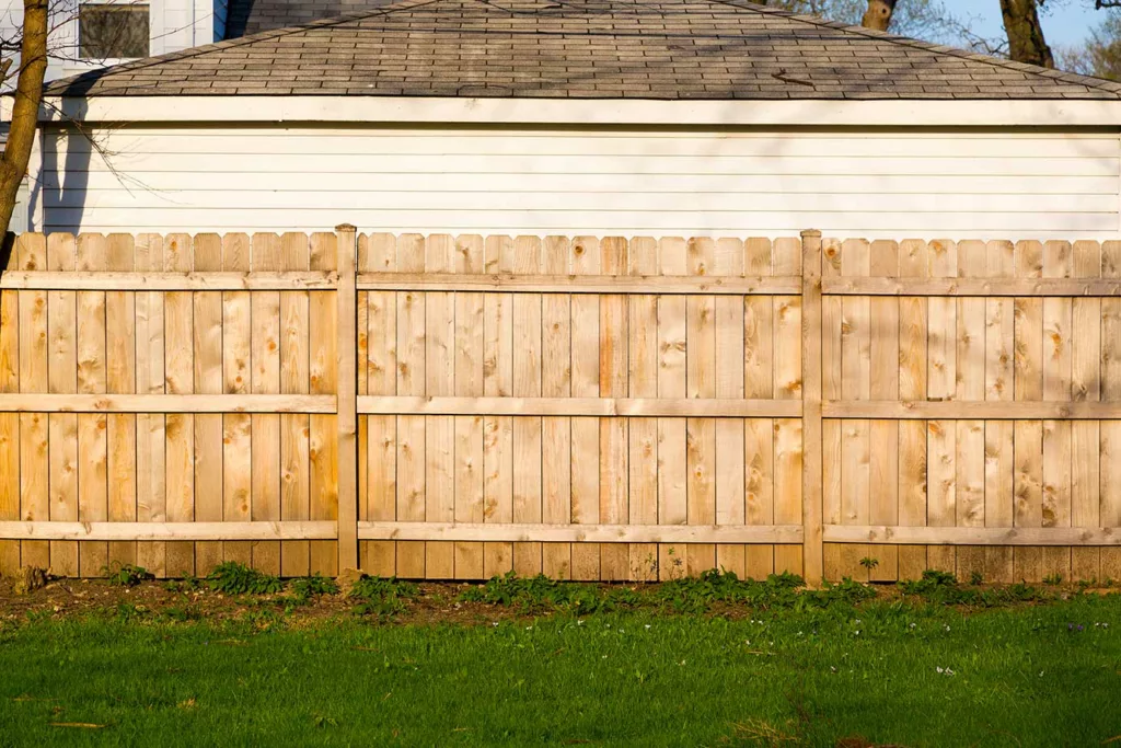 New fence with unpainted or stained boards that touch the ground, with garage wall and roof behind.