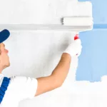 A professional residential painter in a hat paints a wall blue with a paint roller.