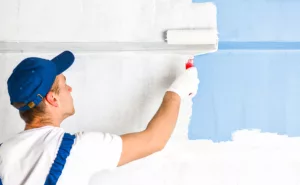 A professional residential painter in a hat paints a wall blue with a paint roller.