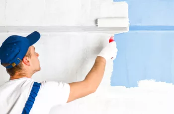 A professional residential painter in a hat paints a wall blue with a paint roller.