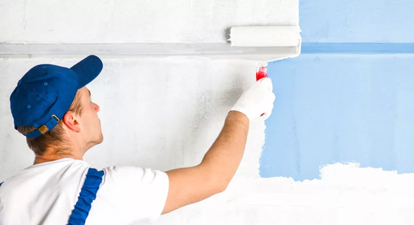 A professional residential painter in a hat paints a wall blue with a paint roller.