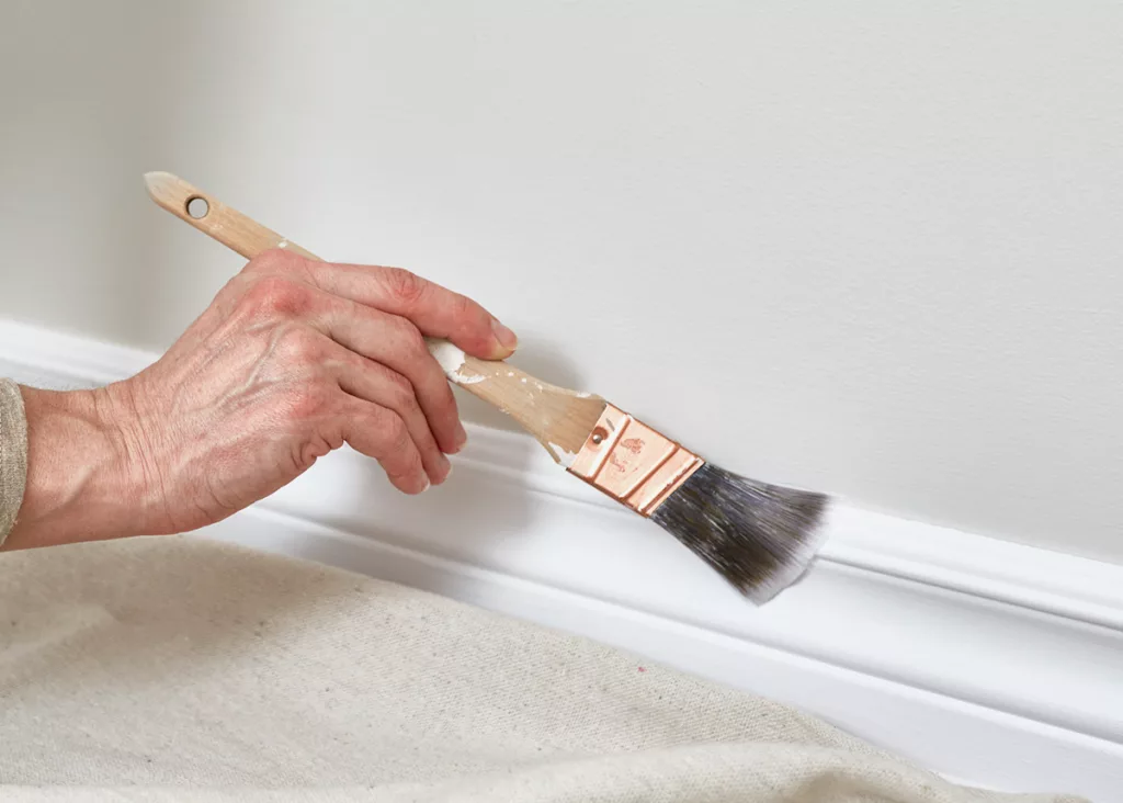 A professional painter repaints interior baseboards with a brush and white paint.