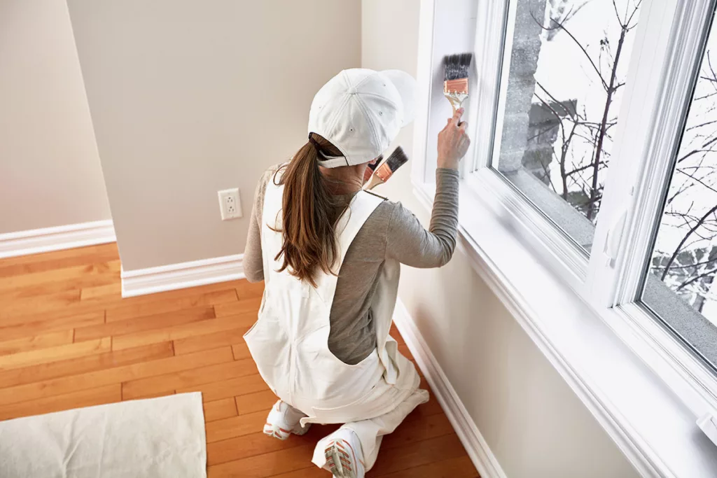 woman in a painter's uniform repainting the interior window trim inside a house