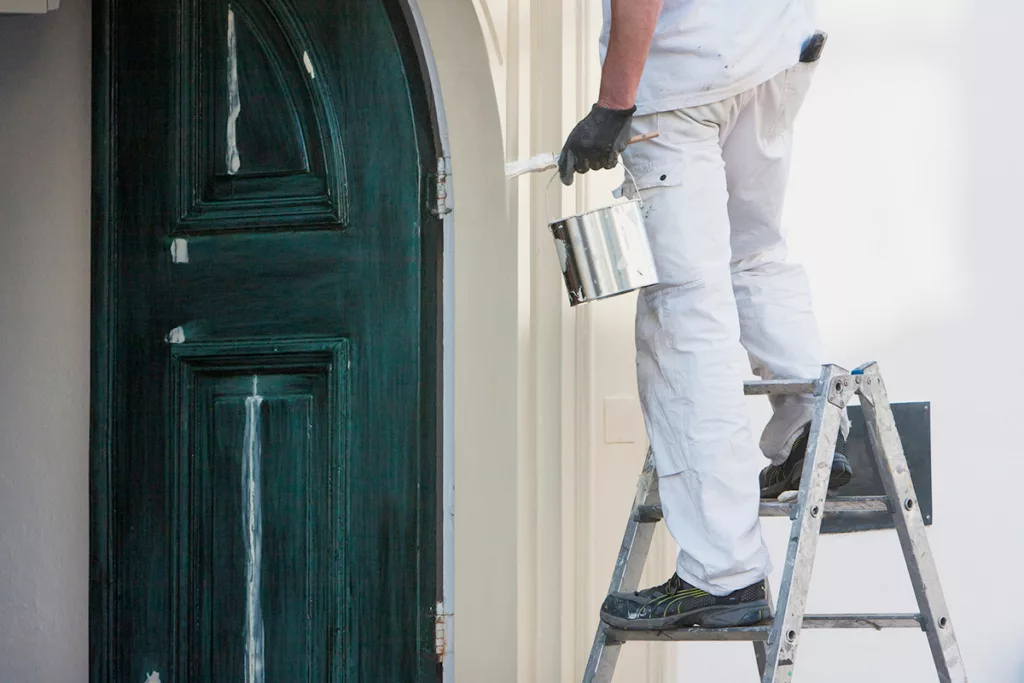 A residential painter stands on a ladder holding a bucket of paint, painting the exterior of a house.