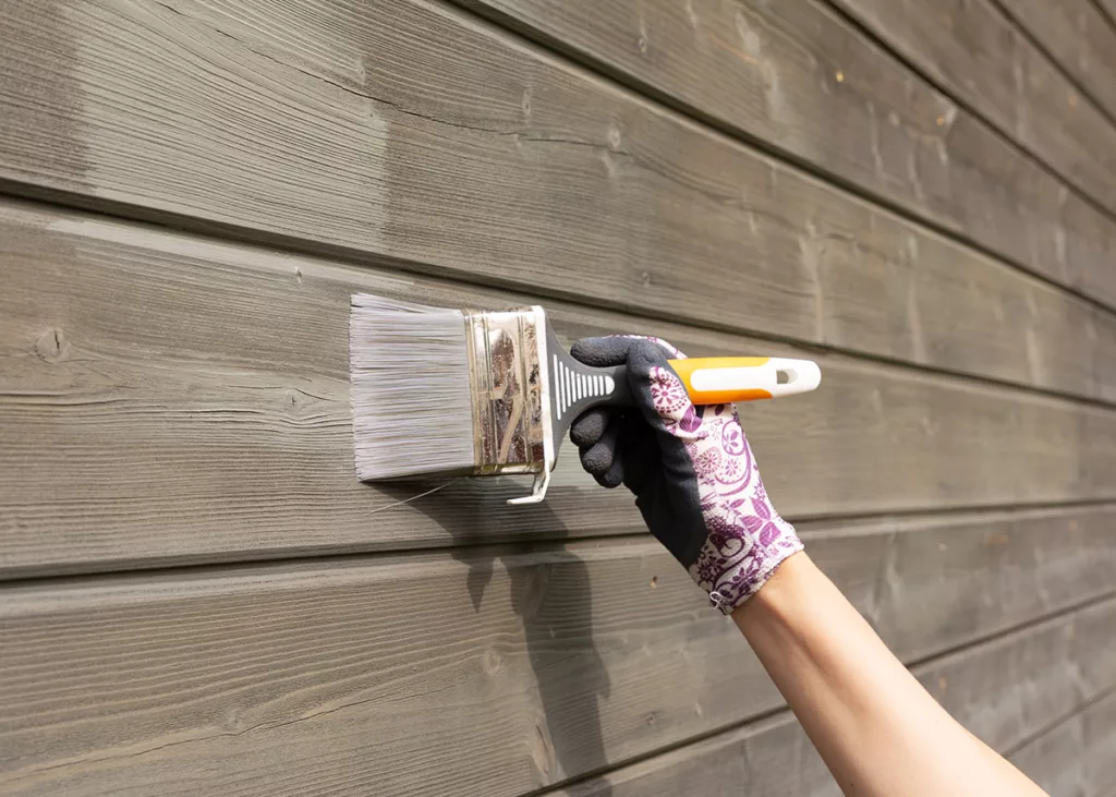 A hand holding a paintbrush paints the exterior wall of a brown house.