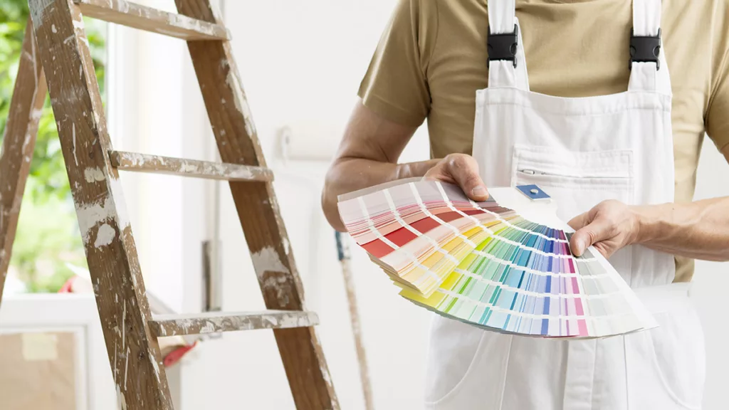 A professional residential painter stands next to a ladder, holding out a selection of paint colors to choose from.