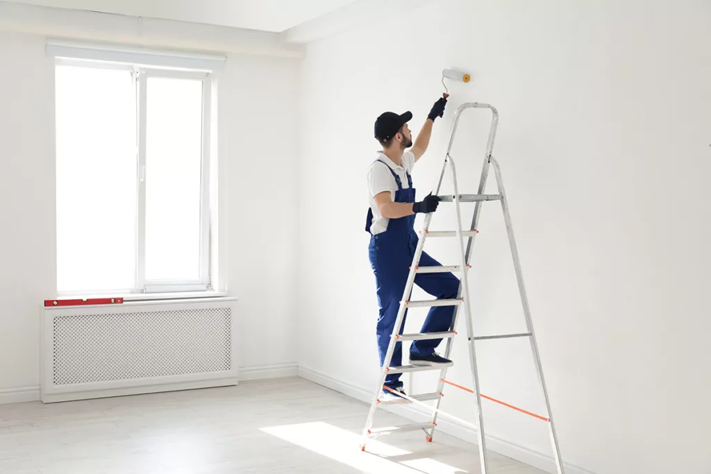 A professional painter stands on a ladder in an empty room, painting the walls white.