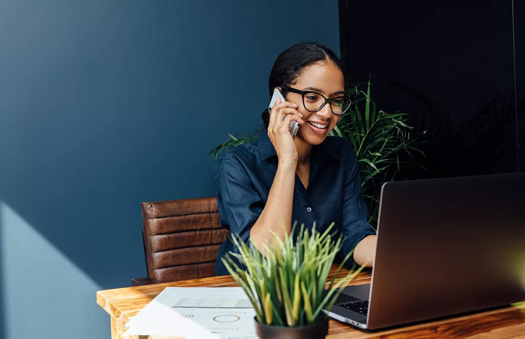 A business woman sits at a desk behind a laptop on the phone in a commercial office with dark blue paint.