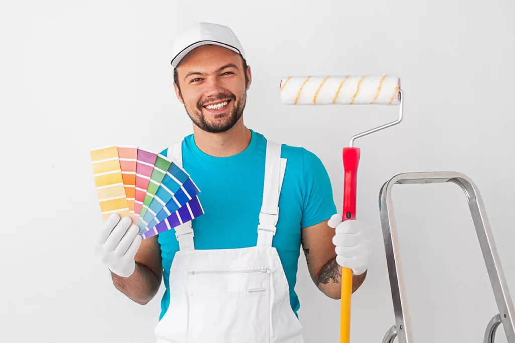 A cheerful professional painter holds a color palette and a roller against a white background next to a step ladder