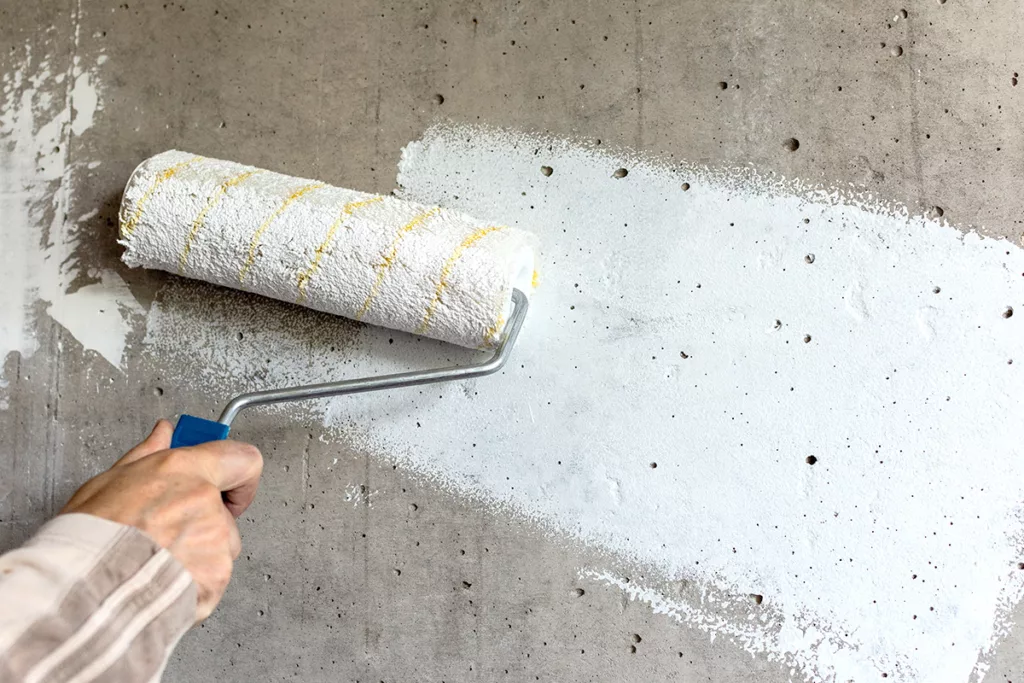 A professional painter uses a paint roller to apply white paint sealer over a dark gray concrete surface.