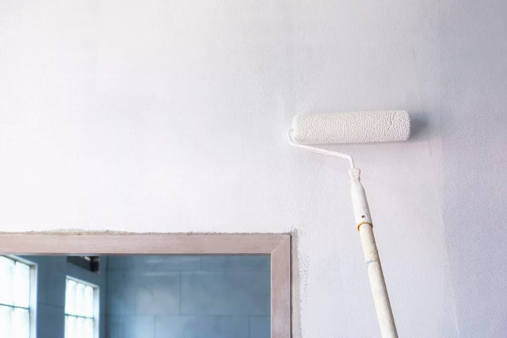 A professional residential painter uses a long paint roller to paint the surface of a wall with white primer.