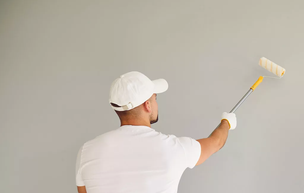 A professional commercial painter wearing white clothes and a white hat paints a wall with a roller.