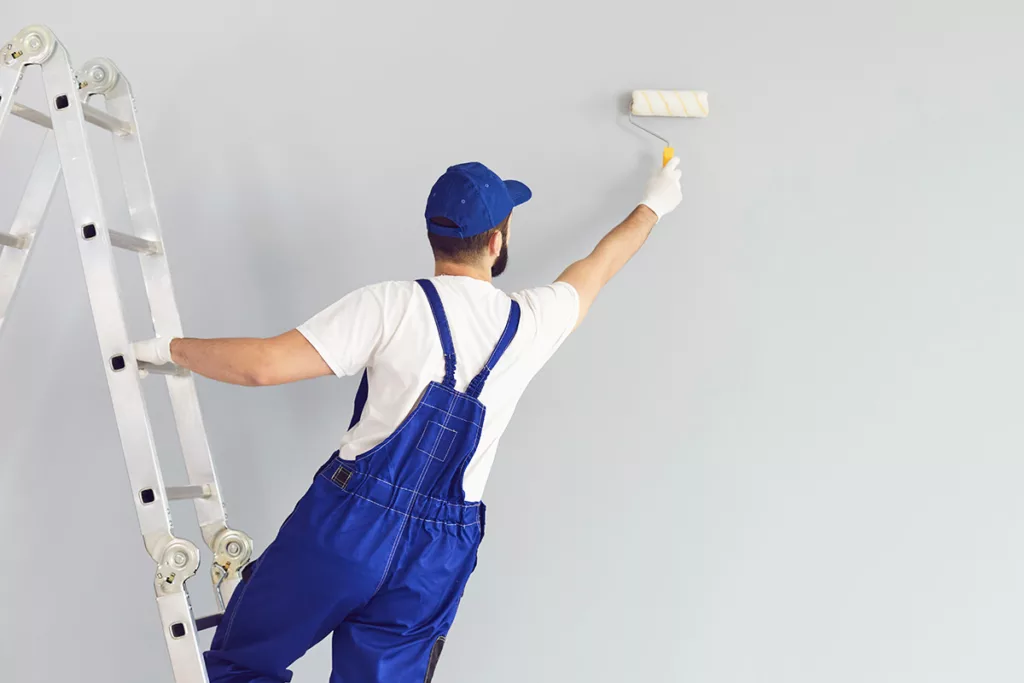A professional painter stands on a ladder and paints a wall gray with a roller for a commercial painting project.