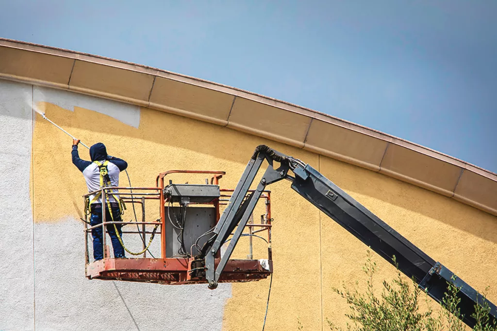 A professional painter stands on a lift, painting the exterior of a commercial building with a sprayer