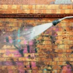 A professional painter pressure rinsing a brick wall to remove graffiti